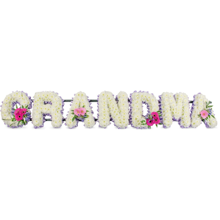 The image shows a beautifully composed funeral tribute spelling the word "GRANDMA" in large, upright letters, each letter made entirely from crisp white chrysanthemums. The chrysanthemums are set densely together, forming a lush, uniform surface that looks soft and cushiony, as if a loving hand has smoothed every bloom into place. Their bright white petals reflect a gentle, even light, giving the whole tribute an air of cleanliness and peace, not unlike the feeling of a clear morning over Chingford's open green spaces. Tracing the outline of each letter is a narrow band of lilac ribbon, its satin sheen catching the light at the edges and lending the tribute a refined, almost regal finish. This subtle purple border helps each letter stand out, ensuring that "GRANDMA" reads clearly even from a distance at a funeral service. Interspersed along the design are three small, round clusters of accent flowers: vivid pink gerberas with detailed centres, soft pink spray roses, and carefully placed sprigs of fresh greenery. These clusters appear on the initial "G," one of the middle "A"s, and the final "A," bringing rhythmic pops of colour and hinting at the personality and warmth of the grandmother being honoured. The green foliage adds texture and a gentle sense of movement, suggesting renewal and continuity. The tribute seems to rest on a discreet white base, ready to be displayed at a local Chingford church, crematorium, or graveside. There is an imagined, subtle fragrance-clean chrysanthemum and light rose-that would mingle quietly with the air, offering a small measure of comfort. This floral "GRANDMA" piece feels like a heartfelt message expressed in petals, crafted with care for a deeply treasured family matriarch.